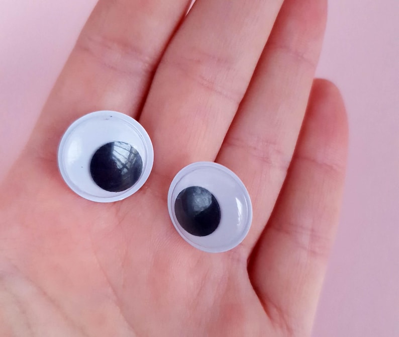 May include: Two white plastic googly eyes with black pupils. The eyes are on a person's hand.