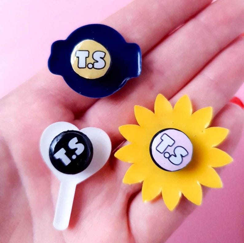 May include: Three different pins with a sunflower, a heart, and a circle design. Each pin has a white background with the letters "T.S" in black.