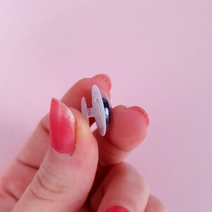 May include: A close-up of a white plastic safety eye with a black pupil. The eye is held between two fingers.