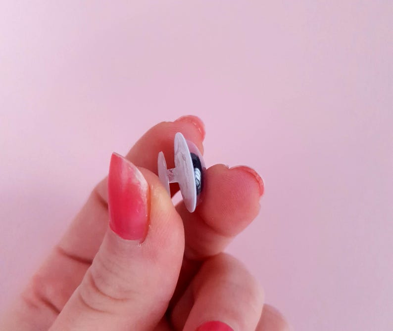 May include: A close-up of a white plastic safety eye with a black pupil. The eye is held between two fingers.