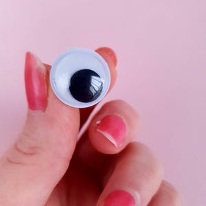 May include: A white plastic googly eye with a black pupil, held by a hand with pink nail polish.