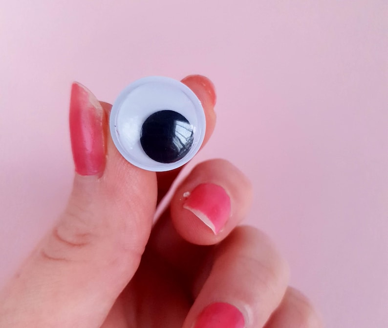 May include: A white plastic googly eye with a black pupil, held by a hand with pink nail polish.