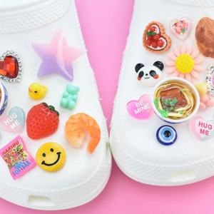 May include: White clogs adorned with a variety of charms. Charms include a star, heart, panda, strawberry, smiley face, and a bowl of noodles. Other charms include a ladybird, gummy bears, and a bag of sweets.