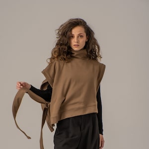 Könnte beinhalten: Eine braune Rollkragen-Pulloverweste mit einer langen Krawatte, die um die Taille gebunden werden kann. Die Weste hat eine lockere Passform und ist aus einem weichen, komfortablen Stoff gefertigt. Das Model trägt eine schwarze Hose und schwarze Stiefel.