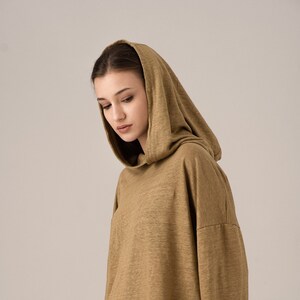 K&ouml;nnte beinhalten: Ein olivgr&uuml;nes, kurz geschnittenes Kapuzensweatshirt. Das lang&auml;rmlige Oberteil hat eine lockere Passform und einen Kordelzug am Saum. Das Model tr&auml;gt eine schwarze Hose.