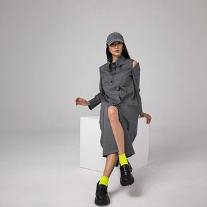 May include: A woman wearing a gray collared dress with a single shoulder cut-out, a gray baseball cap, and bright yellow socks. She is sitting on a white cube.