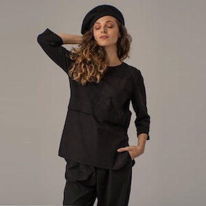 May include: A black, long-sleeved shirt with a layered design and a single pocket. The shirt is worn with black pants and a black beret.