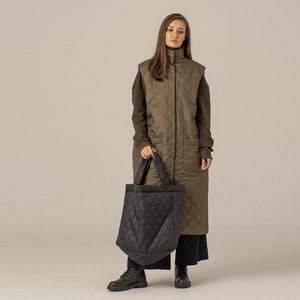 Può includere: Una donna indossa un lungo gilet trapuntato verde oliva con una camicia nera a maniche lunghe sotto. Tiene una borsa tote trapuntata nera.
