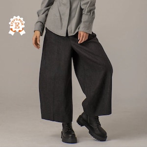 May include: A pair of wide-leg, dark gray pants with a slightly flared hem. The pants are made of a soft, textured fabric and have a relaxed fit. The model is wearing the pants with a light gray button-down shirt and black boots.
