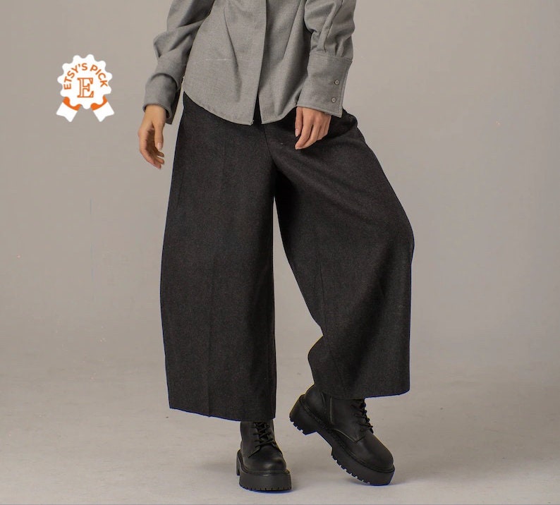May include: A pair of wide-leg, dark gray pants with a slightly flared hem. The pants are made of a soft, textured fabric and have a relaxed fit. The model is wearing the pants with a light gray button-down shirt and black boots.