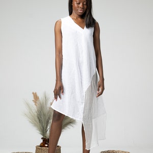 May include: A woman wearing a white sleeveless dress with a draped asymmetrical hem. She is standing on a white background with two wicker baskets and a potted plant.