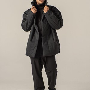 May include: A black quilted jacket with a high collar and a black bucket hat. The person is wearing black pants and black sneakers.