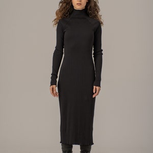Puede incluir: Un vestido midi negro de canal&eacute; con cuello alto y mangas largas. El vestido es ajustado y tiene una falda ligeramente acampanada. La modelo lleva botas de combate negras.