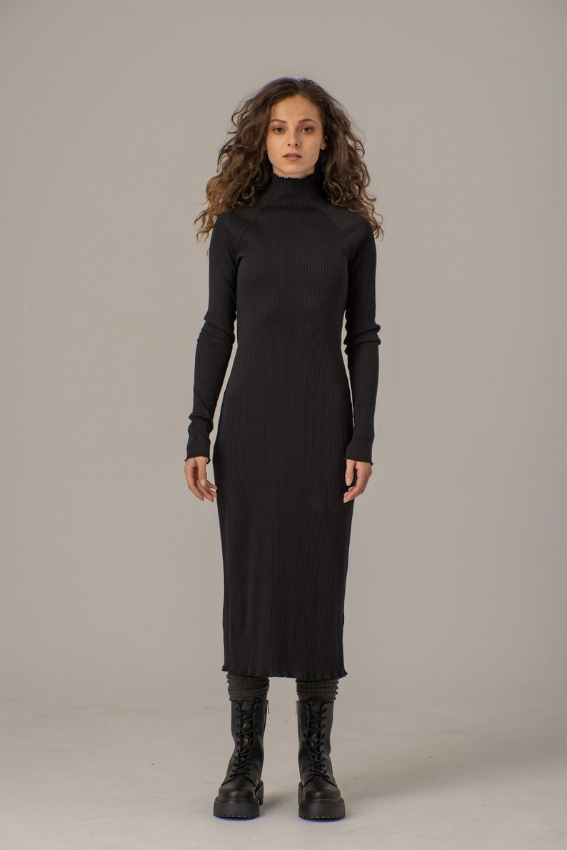 Puede incluir: Un vestido midi negro de canal&eacute; con cuello alto y mangas largas. El vestido es ajustado y tiene una falda ligeramente acampanada. La modelo lleva botas de combate negras.