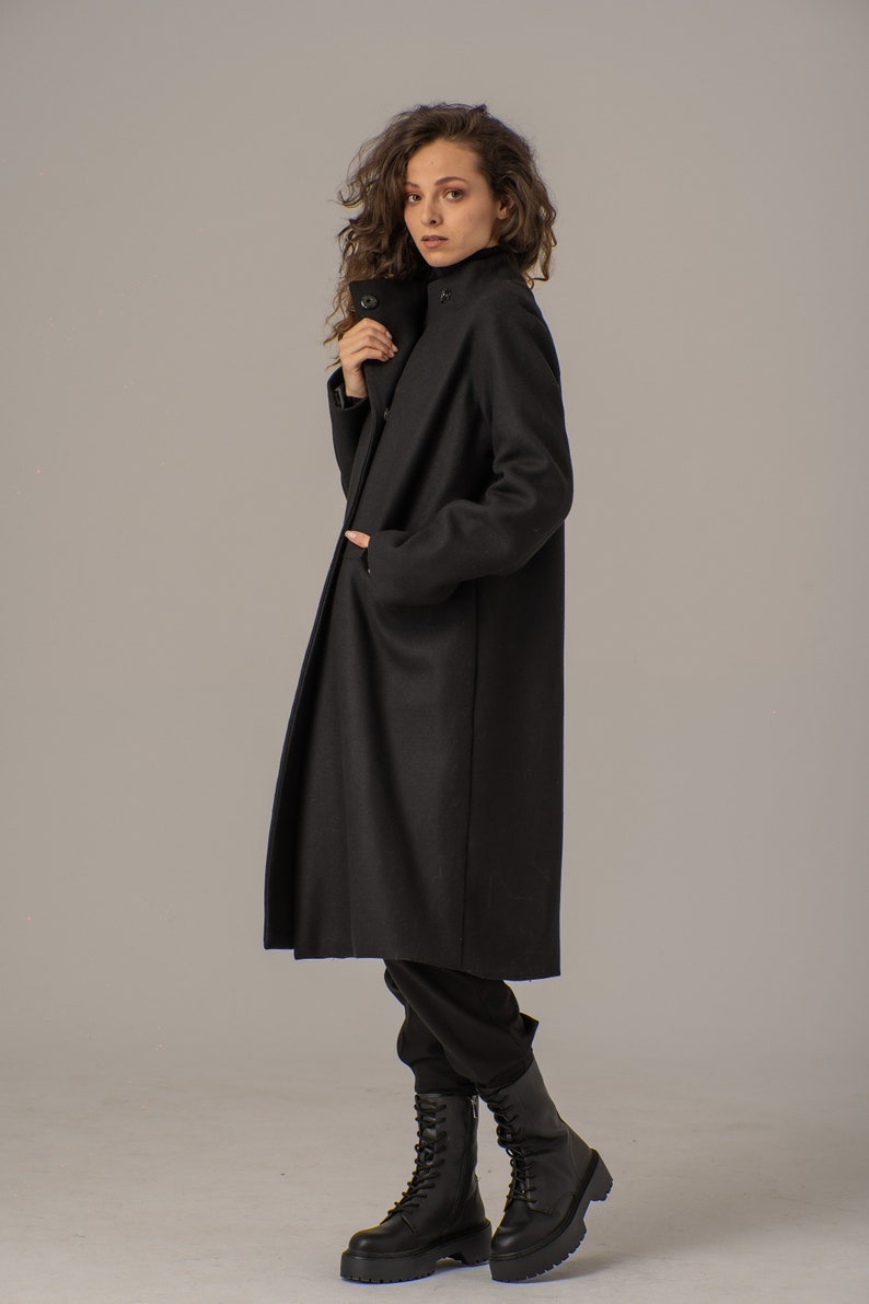 May include: A black long-sleeved coat with a high collar and a button closure. The coat is worn by a person with curly brown hair and is standing on a light gray background.