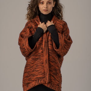 Op de afbeelding: Een vrouw draagt een langarmige, oranje en zwarte gebreide cardigan met een zwarte turtleneck eronder. De cardigan heeft een losse pasvorm en een licht getailleerde lengte.