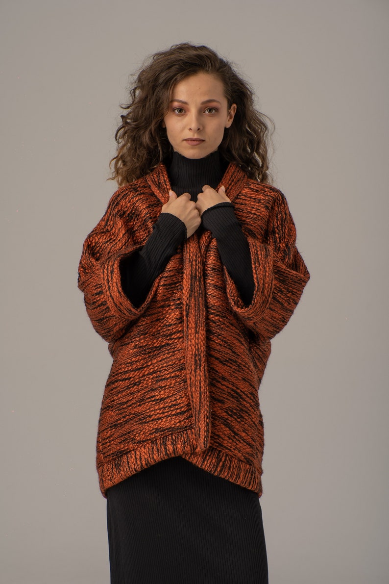 Op de afbeelding: Een vrouw draagt een langarmige, oranje en zwarte gebreide cardigan met een zwarte turtleneck eronder. De cardigan heeft een losse pasvorm en een licht getailleerde lengte.