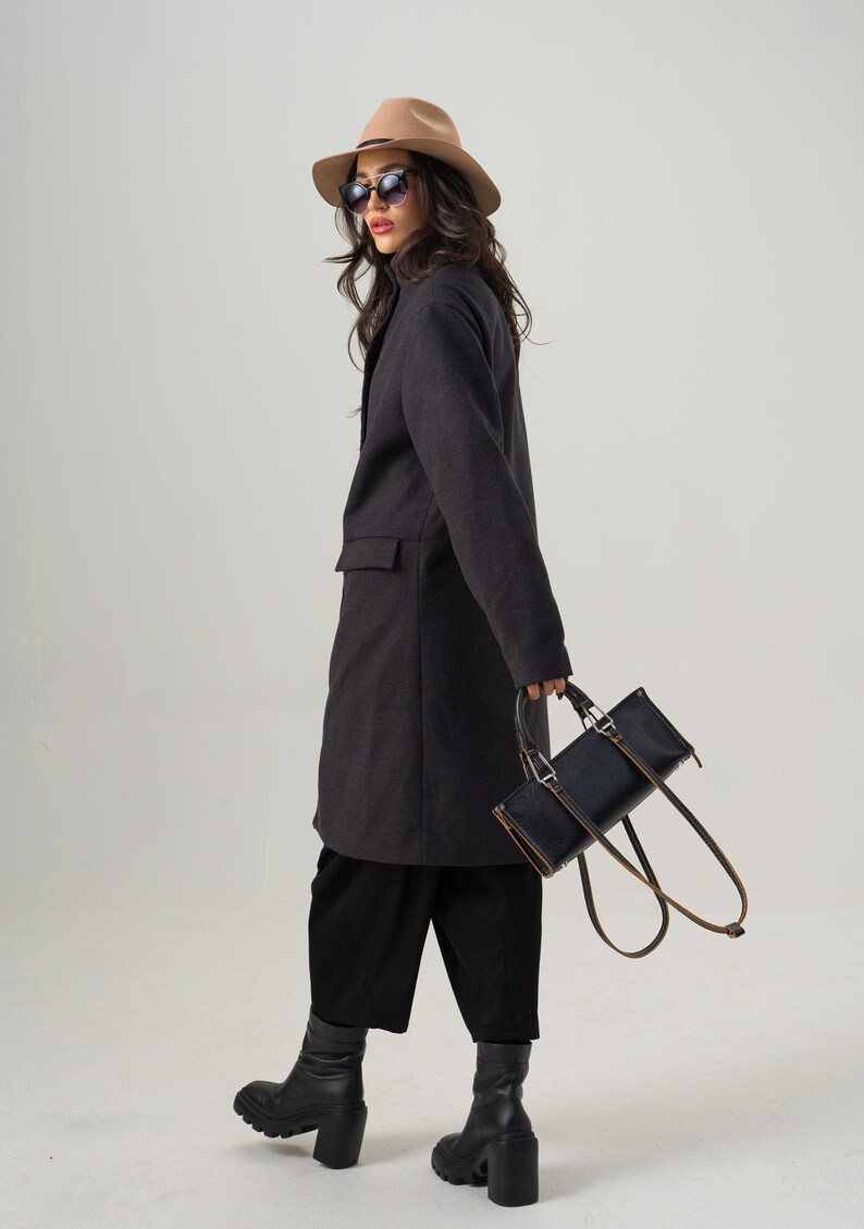 May include: A dark gray coat with a tailored fit, paired with black culottes and chunky black boots. A tan fedora and sunglasses complete the look. A black rectangular handbag with brown straps is held in the hand.