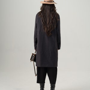 May include: A person wearing a dark gray long coat, black pants, and black boots, holding a small black purse with a brown strap. They are also wearing a tan fedora hat with a black band.