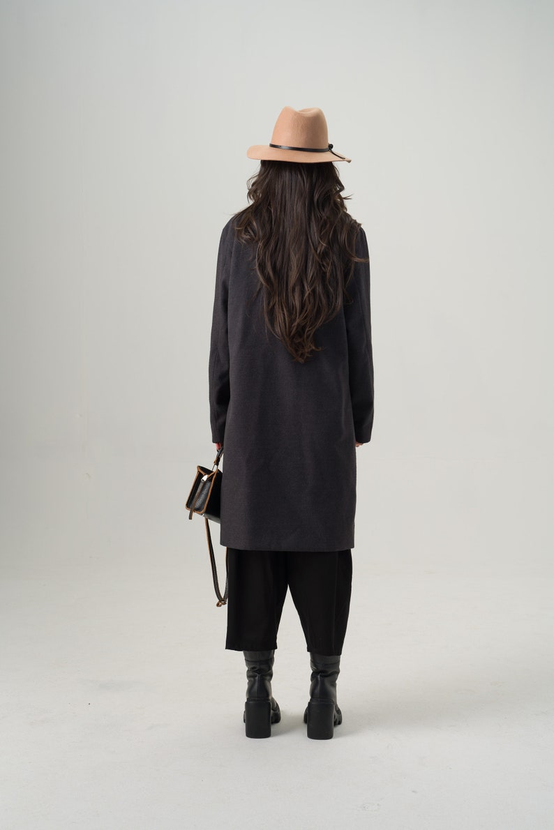 May include: A person wearing a dark gray long coat, black pants, and black boots, holding a small black purse with a brown strap. They are also wearing a tan fedora hat with a black band.