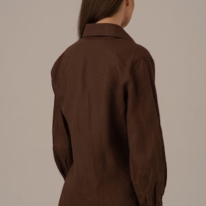 May include: A brown linen shirt with long sleeves and a button-up collar. The shirt has a relaxed fit and a slightly gathered look at the shoulders.