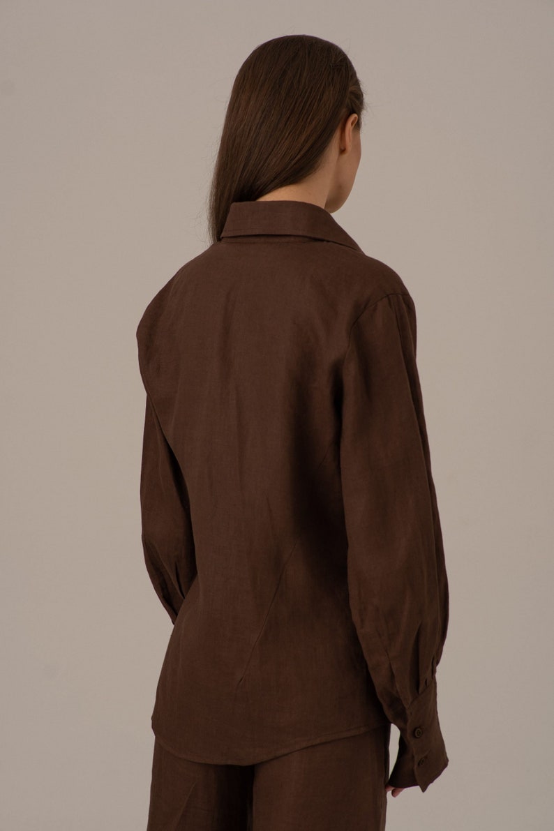 May include: A brown linen shirt with long sleeves and a button-up collar. The shirt has a relaxed fit and a slightly gathered look at the shoulders.