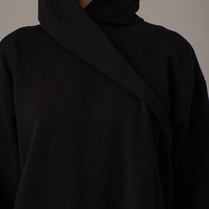 Pode incluir: Uma camisola de capuz preta com um capuz longo e drapeado. O capuz est&aacute; dobrado sobre si mesmo e o tecido &eacute; suave e liso.