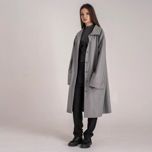May include: A gray long-sleeved coat with a button closure and large pockets. The coat is worn over a black top and black pants.