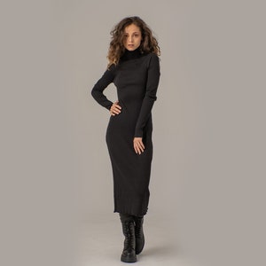 Puede incluir: Una mujer con un vestido largo de cuello alto negro con textura de canal&eacute;. Est&aacute; de pie con la mano en la cadera y lleva botas negras de tobillo.
