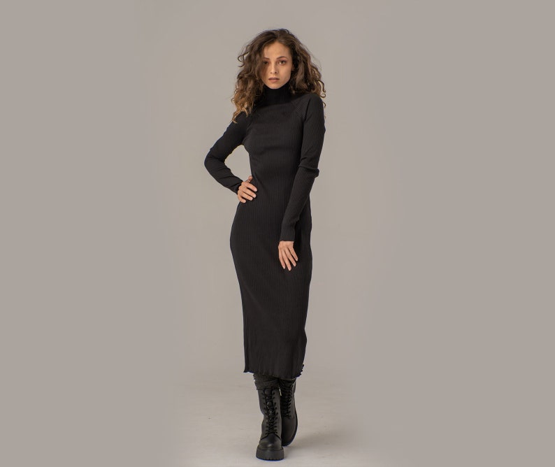 Puede incluir: Una mujer con un vestido largo de cuello alto negro con textura de canal&eacute;. Est&aacute; de pie con la mano en la cadera y lleva botas negras de tobillo.