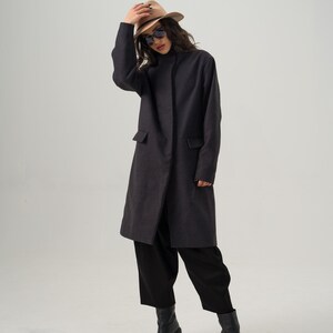 May include: A charcoal gray coat with two front pockets, paired with black pants and boots. The coat is knee-length and has a simple, elegant design. A beige fedora and sunglasses complete the outfit.