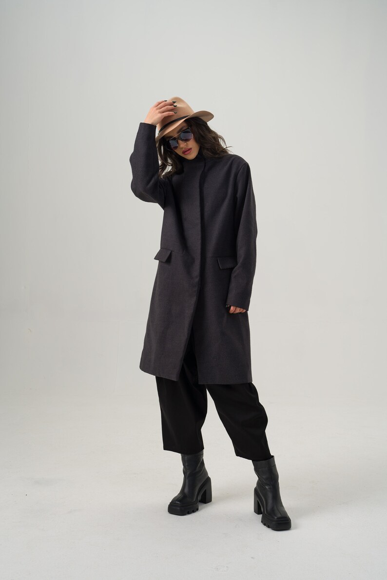 May include: A charcoal gray coat with two front pockets, paired with black pants and boots. The coat is knee-length and has a simple, elegant design. A beige fedora and sunglasses complete the outfit.