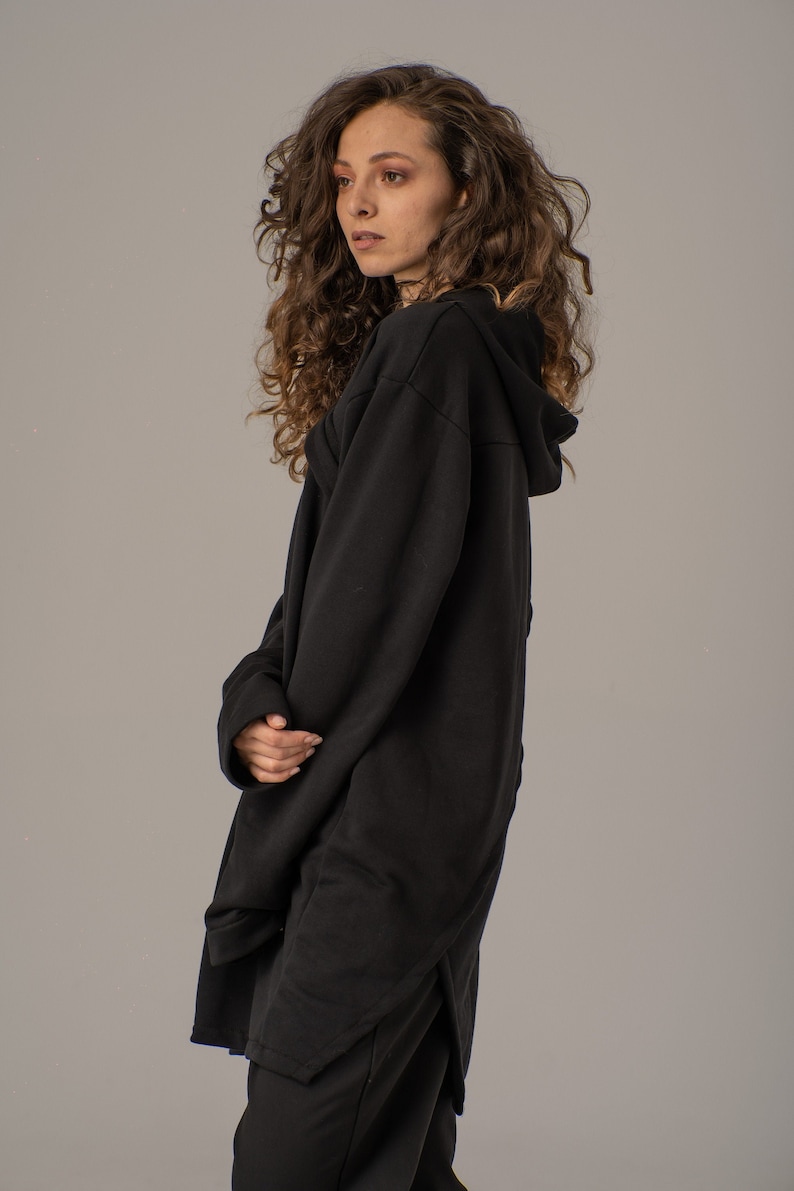 Pode incluir: Uma mulher vestindo um moletom com capuz preto de mangas compridas e bainha longa assim&eacute;trica. Ela tem cabelos longos e cacheados castanhos e est&aacute; olhando para o lado.
