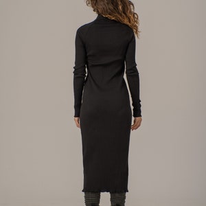 Puede incluir: Un vestido negro de punto de canal&eacute; con cuello alto y mangas largas. El vestido es ajustado y tiene una ligera forma de A. La modelo lleva botas negras.