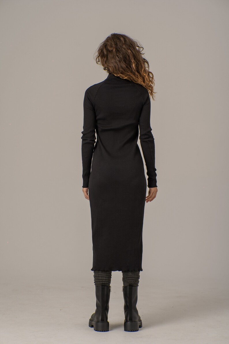 Puede incluir: Un vestido negro de punto de canal&eacute; con cuello alto y mangas largas. El vestido es ajustado y tiene una ligera forma de A. La modelo lleva botas negras.