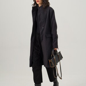 May include: A woman wearing a dark gray coat, black pants, and black boots. She is also wearing a tan fedora hat and sunglasses. She is carrying a black handbag with brown accents.