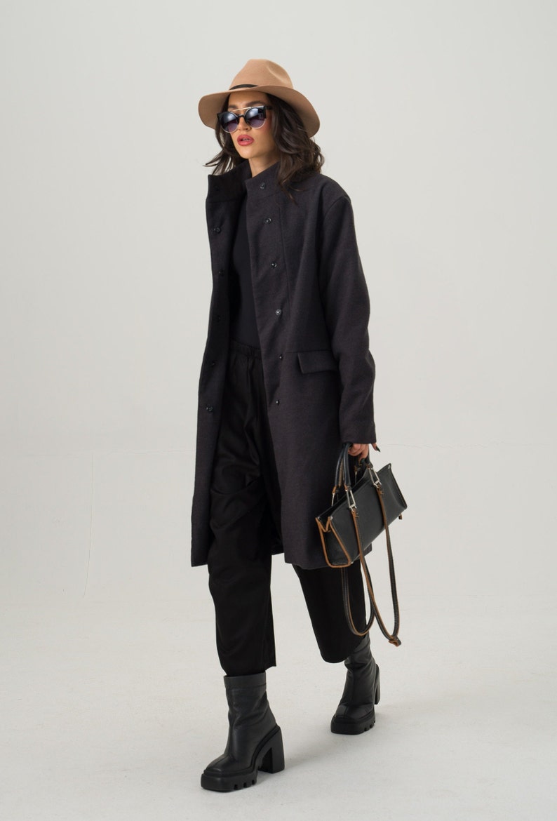 May include: A woman wearing a dark gray coat, black pants, and black boots. She is also wearing a tan fedora hat and sunglasses. She is carrying a black handbag with brown accents.