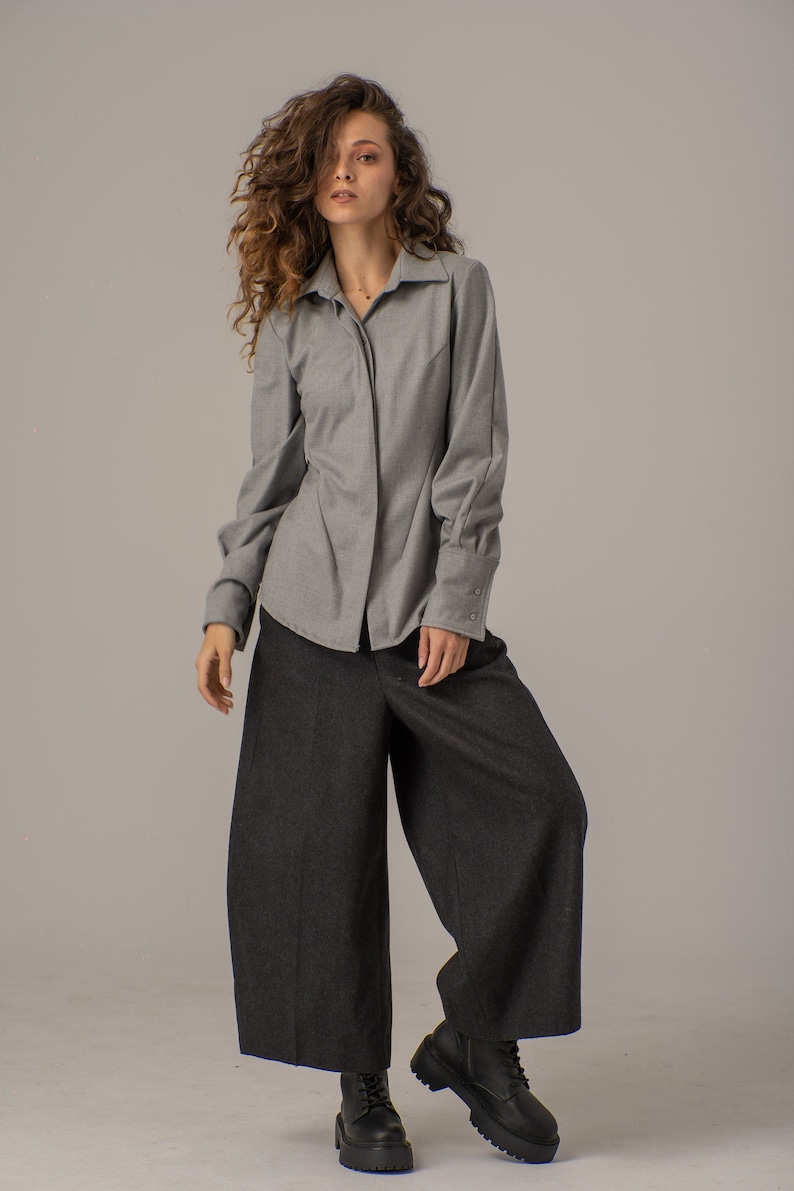 May include: A woman wearing a gray collared shirt with long sleeves and black wide-leg pants. She is wearing black lace-up boots.