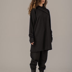 Pode incluir: Um moletom preto de manga comprida com capuz, com um caimento solto e uma silhueta longa e fluida. A modelo est&aacute; usando cal&ccedil;as pretas e botas pretas de combate.
