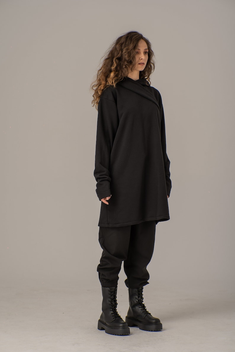 Pode incluir: Um moletom preto de manga comprida com capuz, com um caimento solto e uma silhueta longa e fluida. A modelo est&aacute; usando cal&ccedil;as pretas e botas pretas de combate.