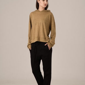 K&ouml;nnte beinhalten: Ein l&auml;ssiges Outfit mit einem beigefarbenen Kapuzenpullover und einer schwarzen Hose. Der lang&auml;rmlige Kapuzenpullover hat eine Kapuze mit Kordelzug und wird mit einer locker sitzenden schwarzen Hose kombiniert. Schwarze Schuhe vervollst&auml;ndigen das Ensemble.
