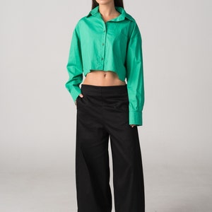 Op de afbeelding: Een vrouw draagt een groen getailleerd shirt en een zwarte wijde broek. Ze staat met haar handen in haar zakken.
