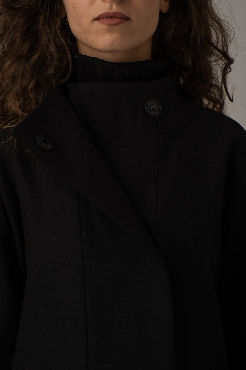 May include: A black mock neck coat with a single button closure. The coat has a long, flowing silhouette and a textured fabric.