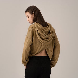 K&ouml;nnte beinhalten: Ein beigefarbener Kapuzenpullover mit langen &Auml;rmeln und einem einzigartigen offenen R&uuml;ckendesign. Der Hoodie hat eine drapierte Kapuze und wird mit einer schwarzen Hose kombiniert. Der Stoff wirkt weich und bequem, geeignet f&uuml;r Freizeitkleidung.