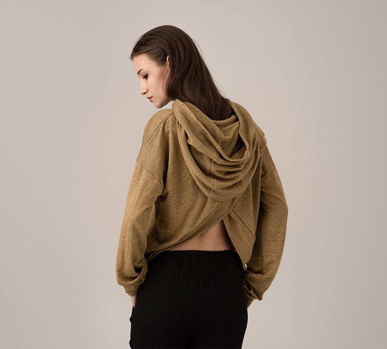 K&ouml;nnte beinhalten: Ein beigefarbener Kapuzenpullover mit langen &Auml;rmeln und einem einzigartigen offenen R&uuml;ckendesign. Der Hoodie hat eine drapierte Kapuze und wird mit einer schwarzen Hose kombiniert. Der Stoff wirkt weich und bequem, geeignet f&uuml;r Freizeitkleidung.