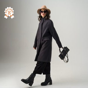May include: A charcoal gray coat with a high collar and a single-breasted design. The coat is paired with black pants and chunky black boots. A black handbag is carried in the hand. The person is wearing a tan hat and sunglasses.