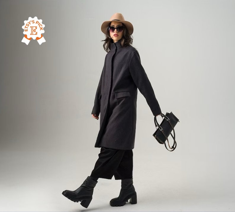 May include: A charcoal gray coat with a high collar and a single-breasted design. The coat is paired with black pants and chunky black boots. A black handbag is carried in the hand. The person is wearing a tan hat and sunglasses.