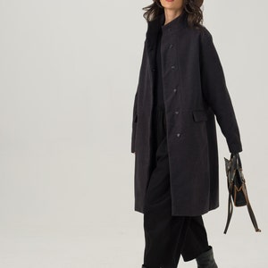 May include: A dark gray, double-breasted coat with a stand-up collar. The coat has two front pockets and is worn with black pants and boots. A tan hat and sunglasses complete the look. A black handbag with brown accents is held in the hand.