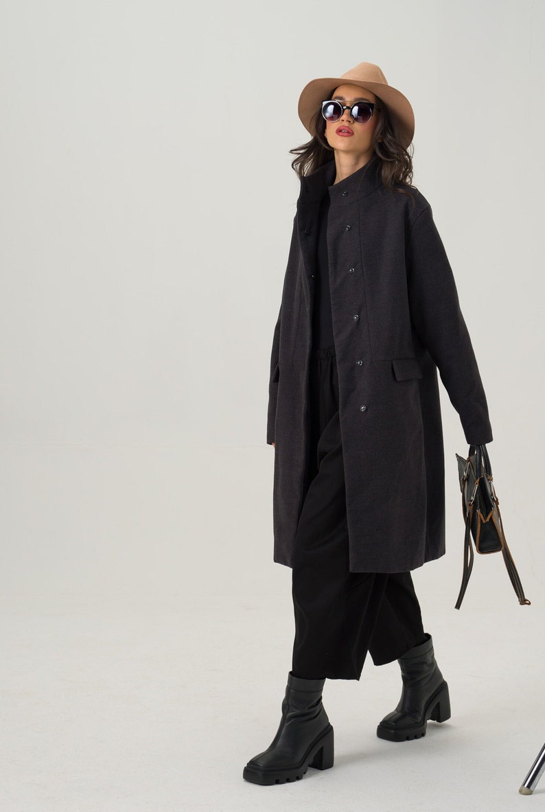 May include: A dark gray, double-breasted coat with a stand-up collar. The coat has two front pockets and is worn with black pants and boots. A tan hat and sunglasses complete the look. A black handbag with brown accents is held in the hand.
