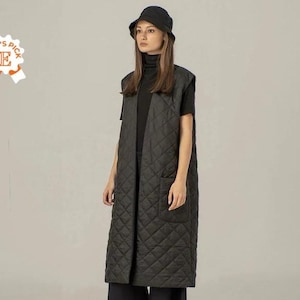 Può includere: Un gilet imbottito nero con un taglio lungo e dritto e una singola tasca sul lato sinistro. Il gilet è indossato sopra un dolcevita nero e un cappello a secchiello nero.